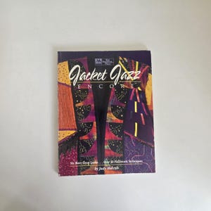 以下が含まれることがあります： Judy Murrub著のソフトカバーの教本『Jacket Jazz Encore』。表紙には、紫、黄色、黒の布地パターンを使用したカラフルなパッチワークジャケットのデザインが描かれています。表紙には「Jacket Jazz Encore」、「Six More Great Looks... Over 30 Patchwork Techniques」、「The Patchwork Place」という文字が記載されています。