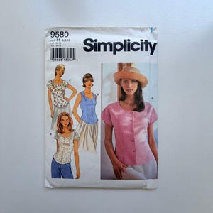 May include: A vintage Simplicity sewing pattern with illustrations of women's tops. The pattern includes a pink button-down shirt, a floral top, and a blue tank top. The pattern number is 9580.