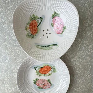 Op de afbeelding: Twee witte keramische borden met een gestructureerd, geribbeld ontwerp. Het grotere bord heeft een unieke vorm en toont illustraties van tomaten, sla en salami. Het kleinere bord heeft een ronde vorm en vergelijkbare voedselillustraties.