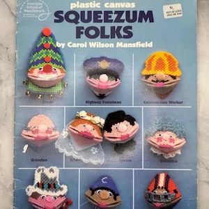 May include: A vintage plastic canvas pattern book titled "Squeezum Folks" by Carol Wilson Mansfield. The cover features colorful, handcrafted figures including a clown, highway patrolman, construction worker, and others. The book is from the American School of Needlework.
