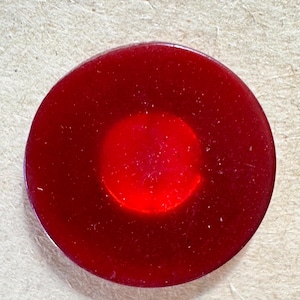 May include: A circular, flat button in a deep, translucent red hue. The button features a smooth, glossy surface with a slightly darker, concentrated red circular centre, creating a subtle gradient effect across the face of the round fastener.