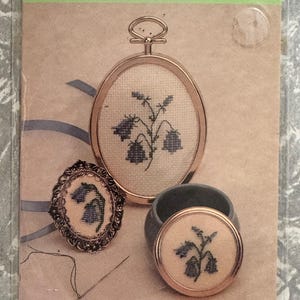 May include: A craft kit featuring a floral design with the text "Harebell". The kit includes an oval frame, a small round frame, and a small blue porcelain box. The design is stitched on 22 count Hardanger fabric.