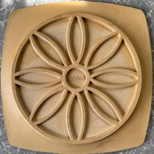 Può includere: Un oggetto quadrato color beige con un motivo floreale circolare al centro. La parola "Ansell" è visibile al centro del motivo floreale. L'oggetto ha una superficie strutturata.