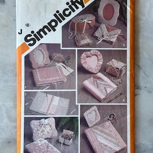 May include: Vintage Simplicity sewing pattern 5296. The pattern features various fabric projects, including a heart-shaped pillow, a small box, and a picture frame. The projects are in shades of pink and white, with lace and ribbon accents. The pattern cover is orange and white.