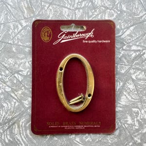 May include: A brass numeral "0" on a maroon card with the "Gainsborough" logo and text. The card also includes the text "SOLID BRASS NUMERALS" and "A PRODUCT OF GAINSBOROUGH HARDWARE INDUSTRIES LIMITED MELBOURNE, AUSTRALIA".