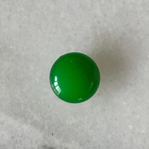 以下が含まれることがあります： 滑らかで光沢のある反射面を持つ、小さな円形の無地の緑色のボタン。この物体は完全に丸く、目に見える上面全体に鮮やかで均一なエメラルドグリーンの色合いが施されています。
