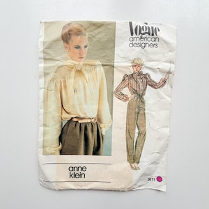 May include: A Vogue American Designers sewing pattern envelope for Anne Klein, number 2611. The front features a photograph of a person wearing a cream-coloured blouse with a tie neck and brown trousers, alongside a fashion illustration of the same outfit.