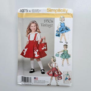 May include: Simplicity sewing pattern 1075 for a 1950s vintage style pinafore dress and matching tote bag. The pattern envelope features illustrations of a red pinafore with white poodle appliqués, a blue pinafore, and a green plaid pinafore design.