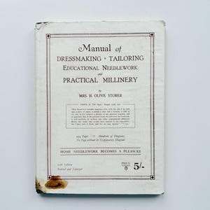 May include: A vintage instructional book titled Manual of Dressmaking, Tailoring, Educational Needlework and Practical Millinery by Mrs. H. Olive Storer. The off-white cover features dark brown text, a decorative border, and a visible stain at the bottom.