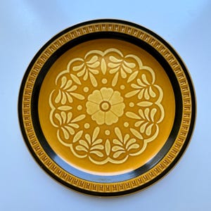 May include: A round ceramic plate featuring a mustard yellow center with a raised, lighter yellow floral and leaf mandala pattern. The wide rim is dark brown with a repeating geometric border of small, raised rectangular indentations.