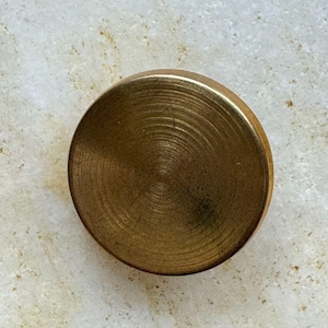 May include: A circular brass button featuring a brushed metallic finish with visible concentric circular texture lines across the surface. The object has a smooth, rounded edge and a flat top profile, presenting a consistent golden-brown tone.