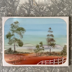 May include: A rectangular painting of a landscape with a blue sky and a red-brown foreground. Trees with green foliage are scattered across the scene, and a white fence runs along the bottom. The painting is signed in the lower left corner.