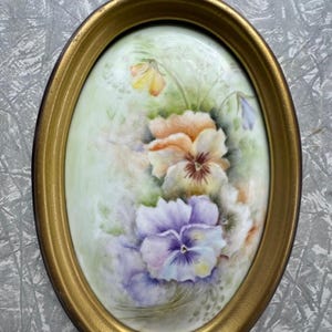 May include: An oval wall plaque featuring a hand-painted porcelain insert with a floral arrangement of pansies in shades of purple, orange, and yellow. The artwork is set within a metallic gold-toned frame with a small hanging loop at the top.