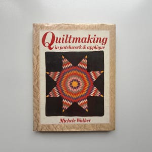 May include: A book titled "Quiltmaking in patchwork & appliqué" by Michele Walker. The cover displays a vibrant starburst pattern in red, orange, yellow, and purple on a black backdrop, framed by a cream-coloured border.