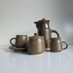 May include: A brown ceramic tea set featuring a tall teapot with a handle, a lidded sugar bowl, a creamer, and a teacup on a saucer. Each piece displays a consistent horizontal ribbed texture and a smooth, dark brown glazed finish throughout.