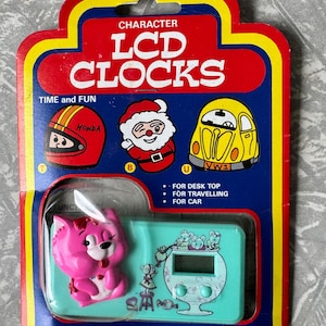 May include: A vintage, unopened package of Character LCD Clocks. The packaging is red, yellow, and blue, with illustrations of a Honda helmet, Santa Claus, and a yellow car. The clock itself is teal with a pink cat figurine.