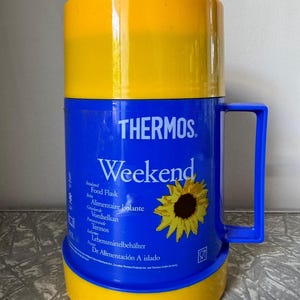 May include: A blue and yellow Thermos food flask with a handle. The flask has the word "Weekend" and a sunflower graphic. The text "THERMOS" is printed on the flask.