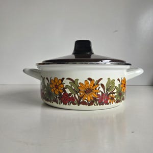 May include: A white enamelled metal cooking pot with two side handles and a dark brown lid. The exterior features a repeating floral pattern in shades of orange, yellow, olive green, and dark red, depicting stylised daisies and small clusters of berries.
