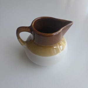 May include: A small ceramic creamer jug featuring a rounded body with a tri-colour glaze. The top rim and handle are dark brown, transitioning to a band of mustard yellow in the middle, and a solid white base at the bottom with a pouring spout.