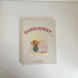 May include: A hardcover book titled Embroidery by Ksynia Marko with a light beige cover. The front features a central illustration of a green vase filled with colorful flowers, red scissors, and the text Including over 60 stitches and many original projects.