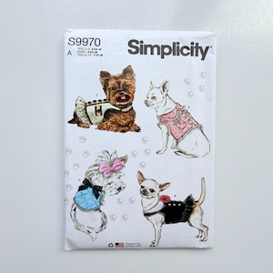 Op de afbeelding: Een Simplicity S9970 naaipatroon-envelop voor hondenkleding in de maten XXS-M. De voorzijde toont illustraties van vier honden in verschillende outfits, waaronder een beige vest, een roze tweed jasje, een blauwe doorgestikte jas en een zwarte jurk.