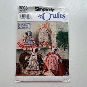 May include: Simplicity Crafts sewing pattern featuring doll designs. The pattern includes instructions for creating fabric dolls with animal-themed faces, including a pig and cows. The dolls are dressed in various outfits, with detailed lace and patterned fabrics.