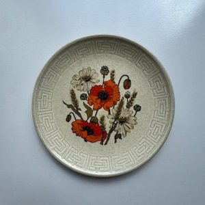 Op de afbeelding: Een ronde keramische bord met een gespikkelde beige afwerking en een centrale bloemenillustratie met feloranje klaprozen, korenaren en witte madeliefjes. De rand is versierd met een herhalend geometrisch Grieks sleutelpatroon in bruintinten.
