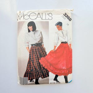 Op de afbeelding: Een McCall's 3268 naaipatroon-envelop met illustraties van twee outfits. De ene toont een witte blouse met ruches en een geruite rok, de andere een witte blouse met ruches en een effen rode rok. Beide zijn voorzien van een bolo-stropdas en een riem.