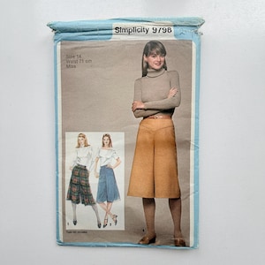 Op de afbeelding: Een Simplicity 9798 naaipatroon-envelop voor maat 14, 71 cm tailleomvang. De voorzijde toont een beige coltrui en een beige rok, met extra illustraties van een geruite rok en een blauwe rok. De tekst op de verpakking luidt Simplicity 9798.