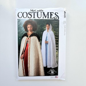 May include: McCall's Costumes sewing pattern M8428 for capes in sizes XS-XL. The envelope features two illustrations: one showing a cream-coloured cape with a black collar over a red dress, and another showing a white hooded cape over a light blue dress.