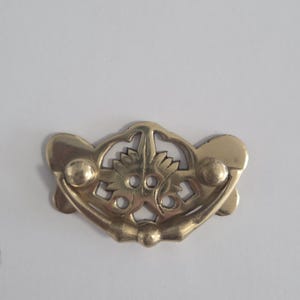 May include: A polished brass drawer pull featuring an ornate, symmetrical openwork design with a central floral motif and a curved, swinging handle attached to two circular mounting points, creating a decorative hardware piece for furniture.