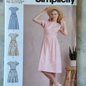 May include: Simplicity sewing pattern S9324. The pattern features illustrations of dresses with pockets. The main image shows a woman wearing a pink and white gingham dress. The pattern is designed and printed in the USA.