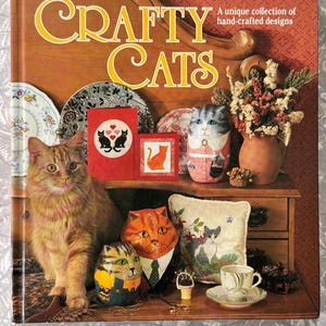May include: A book titled "Crafty Cats" with a collection of hand-crafted designs. The image features a ginger cat, decorative cat figurines, a teacup, and a pillow with cat embroidery. The book and items are arranged on a wooden surface.