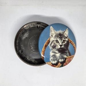 May include: A round metal tin with a removable lid featuring a grey tabby kitten sitting inside a woven basket against a blue background. The interior of the tin is metallic silver with embossed text visible on the bottom surface of the container.