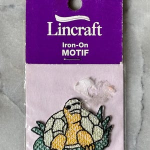 Op de afbeelding: Een Lincraft strijkapplicatie met een geborduurde schildpad met een geel lichaam en een wit schild, omringd door groen blad. De applicatie is bevestigd op een paarse en lichtroze kartonnen achtergrond met een gat om op te hangen.