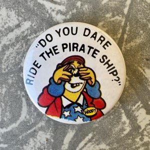 May include: A circular white badge featuring the text "DO YOU DARE RIDE THE PIRATE SHIP?" around a cartoon pirate. The pirate has yellow skin, a red hat, and a blue jacket. The name "Woody" is printed on the badge.