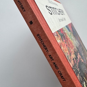 Puede incluir: Un libro antiguo titulado "Stitchery: Art and Craft" de Nile Krevitsky. El libro tiene una cubierta roja con el título y el nombre del autor en negro. La cubierta presenta un diseño abstracto colorido.