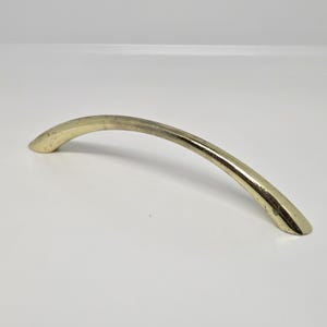 May include: A curved, gold-toned metal cabinet pull handle with a smooth, polished finish. The handle features a simple, arched design that tapers slightly at both ends, showing minor signs of wear and surface texture along its length.