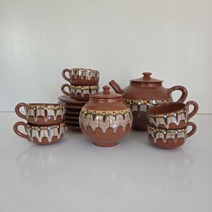 Puede incluir: Un juego de té de cerámica que incluye una tetera, un azucarero con tapa y seis tazas con sus platos a juego. Cada pieza tiene un esmalte marrón cálido con un patrón decorativo de goteo en blanco, verde y marrón oscuro en la parte inferior.