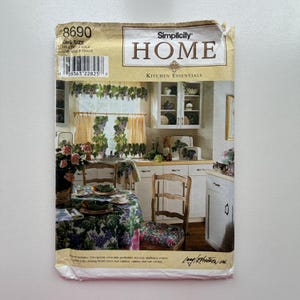 May include: A vintage Simplicity Home Kitchen Essentials sewing pattern. The pattern features a kitchen scene with grape-themed curtains, tablecloth, and chair cushions. The pattern includes aprons, pot holders, tea cosy, appliance covers, and more.