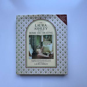 Puede incluir: Un libro de tapa dura titulado "The Laura Ashley Book of Home Decorating". La portada presenta un patrón repetitivo de formas de abanicos verdes y morados. El título del libro está en una fuente decorativa, con "New and Revised Edition".