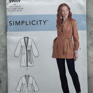 Può includere: Un modello di cucito Simplicity, S9059, con una donna che indossa una giacca marrone e pantaloni neri. Il modello include illustrazioni del design della giacca. Il testo sul modello recita "American Sewing Guild" e "Simplicity".