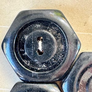 May include: Close-up of several black, hexagonal buttons. Each button has a central, circular indentation with a keyhole-shaped sewing hole. The buttons have a glossy finish and appear to be made of a hard material.