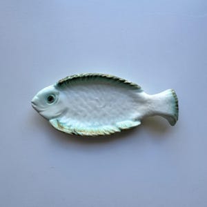 May include: A ceramic dish shaped like a fish with a glossy white finish and subtle light green and brown accents along the edges, fins, and tail. The surface features a textured scale pattern and a defined eye detail on the head of the fish.