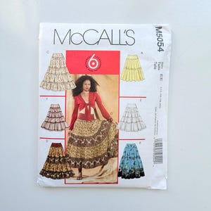 Może przedstawiać: Koperta z wykrojem krawieckim McCall's M5054 na spódnice warstwowe w rozmiarach EE 14-16-18-20. Przód zawiera ilustracje sześciu wariantów spódnic oraz zdjęcie osoby ubranej w długą, wzorzystą spódnicę warstwową i czerwony top typu bolerko.