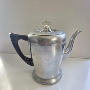 May include: A silver-toned metal coffee percolator featuring a smooth, cylindrical body with a flared base, a curved spout, a black handle, and a removable lid topped with a small, clear glass knob for monitoring the brewing process.