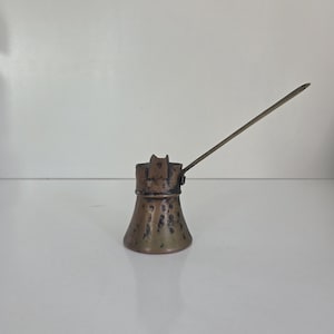 Puede incluir: Una cafetera de cobre desgastada con una base ancha y acampanada, un cuello estrecho y un mango de metal largo y delgado que se extiende diagonalmente hacia arriba. La superficie exterior muestra una pátina oscura y moteada con oxidación.