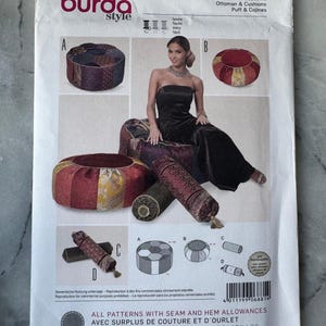 May include: A Burda Style sewing pattern for ottomans and cushions. The cover features illustrations of various ottoman and cushion designs, including round and cylindrical shapes, with a woman in a black dress. The pattern includes seam and hem allowances.