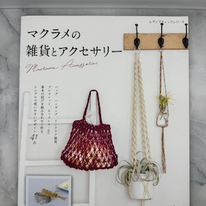 Pode incluir: Um livro japonês intitulado "Macrame Accessories", com uma bolsa de macramê vermelha, um suporte de plantas de macramê branco e um pequeno vaso suspenso de macramê. A capa do livro também mostra uma pequena foto de uma pulseira de macramê.