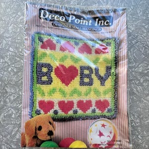 Könnte beinhalten: Ein verpacktes Latch-Hook-Kit von Deco Point Inc. mit dem Text "BABY" und Herzmotiven. Das Kit hat eine fertige Größe von 30 x 30 cm. Ein Plüschwelpe und bunte Bälle sind im Bild zu sehen.
